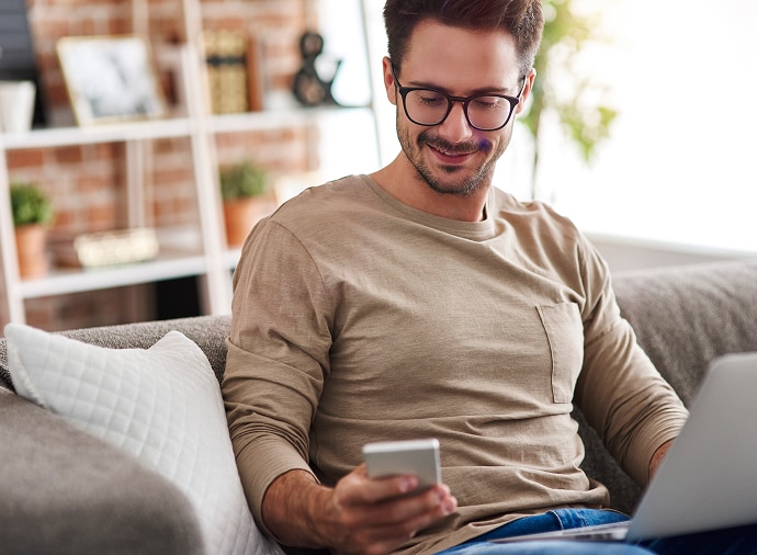 Uomo seduto sul divano con computer portatile sulle ginocchia che guarda il telefono